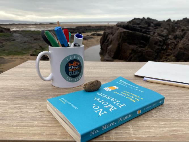 A beachclean branded mug containing pens and a book titled No. More. Plastic. sit on a table overlooking a deserted beach.